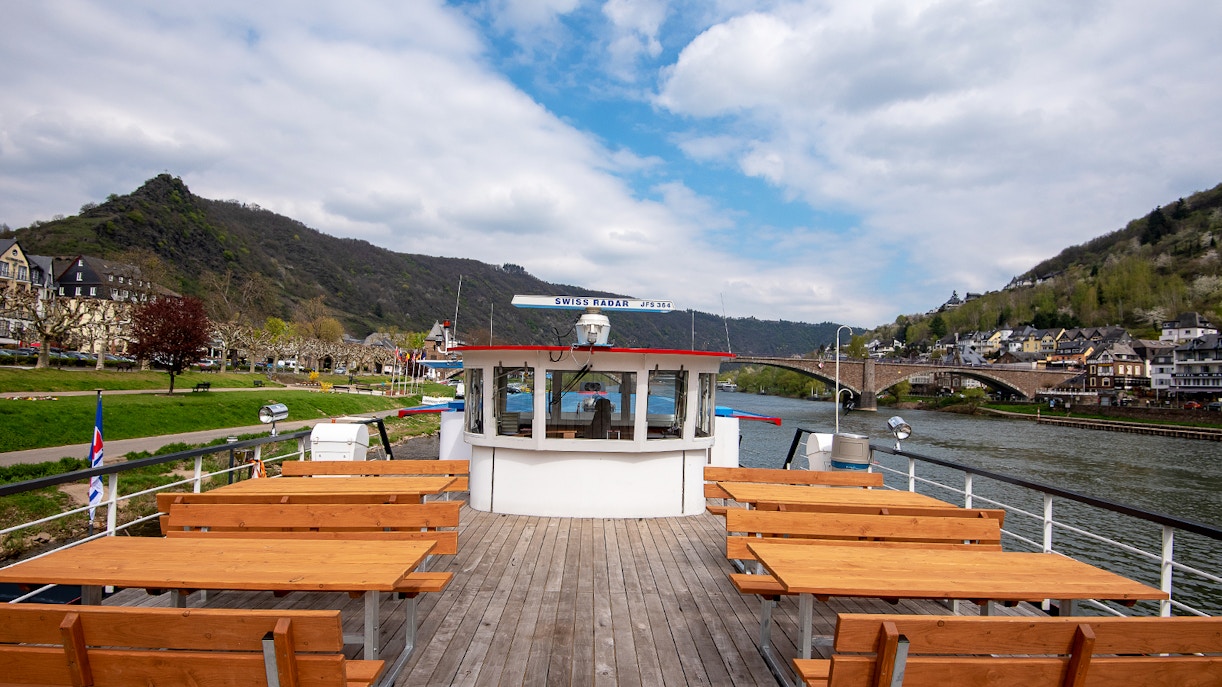 Cruise ship deck with wooden benches on a river, hills and town in the background.