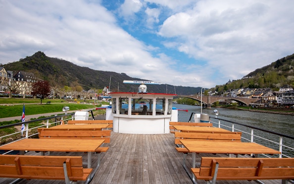 Cruise ship deck with wooden benches on a river, hills and town in the background.