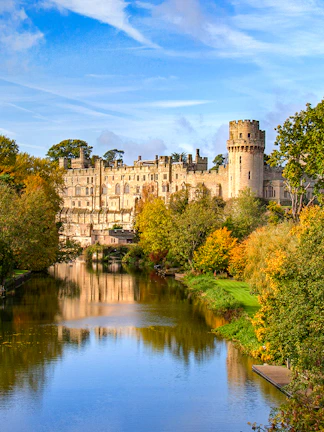 Warwick Castle with river reflection and autumn trees in Warwick, England.
