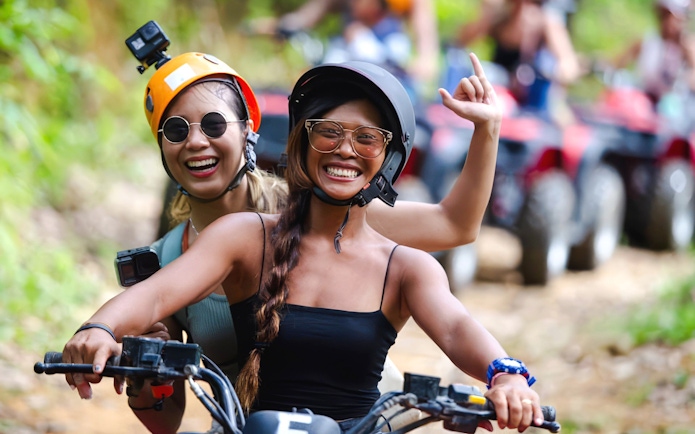 Two girls smiling while riding an ATV through a forest trail.