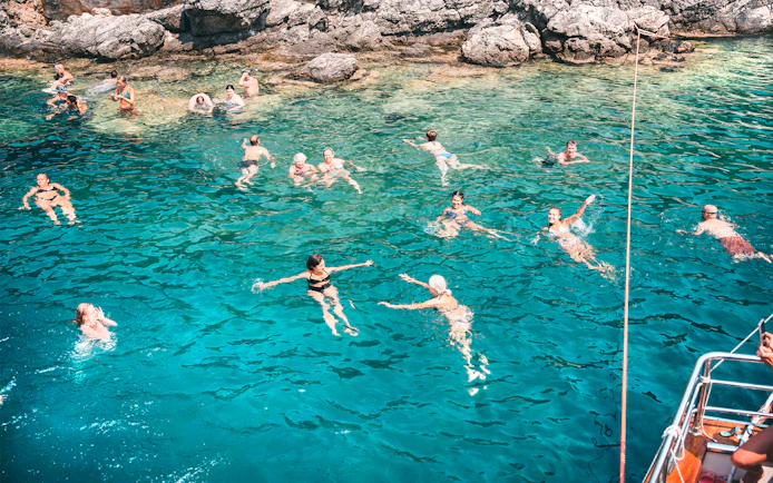 People swimming in clear blue waters near rocky shore during Blue Lagoon & Papanikolis Cruise.