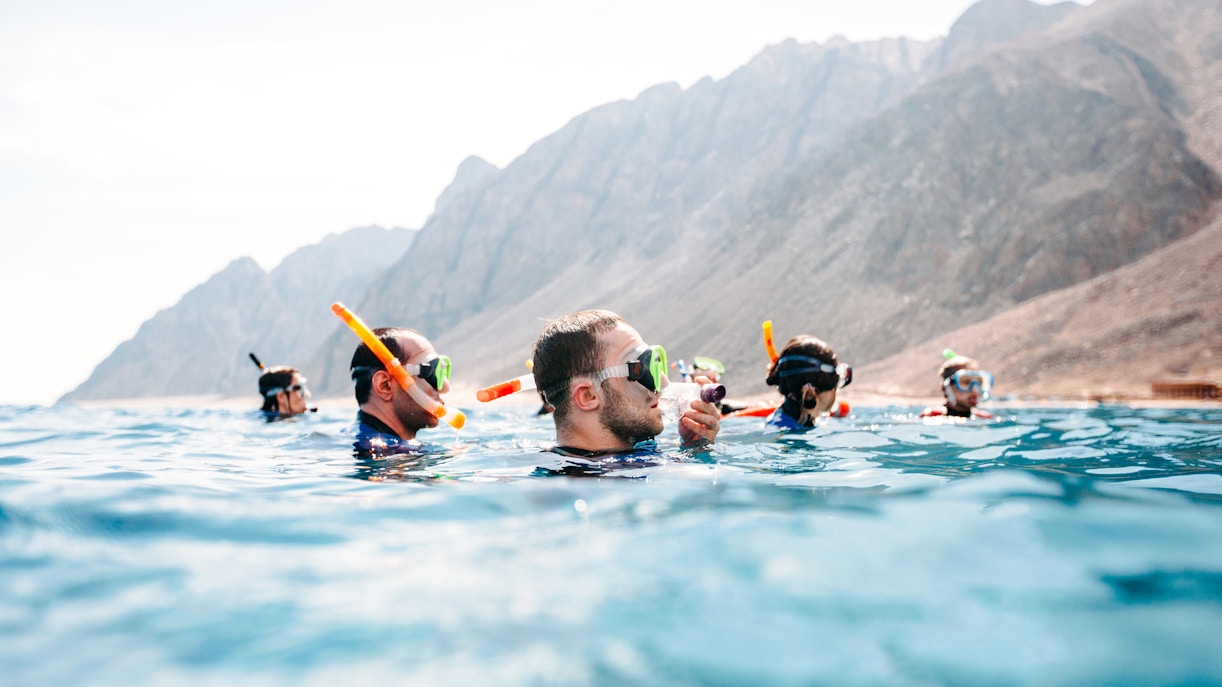 Friends snorkeling in clear sea near rocky coastline.