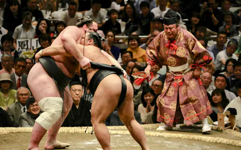 Sumo wrestlers competing in Nagoya Grand Sumo Tournament with referee and audience.