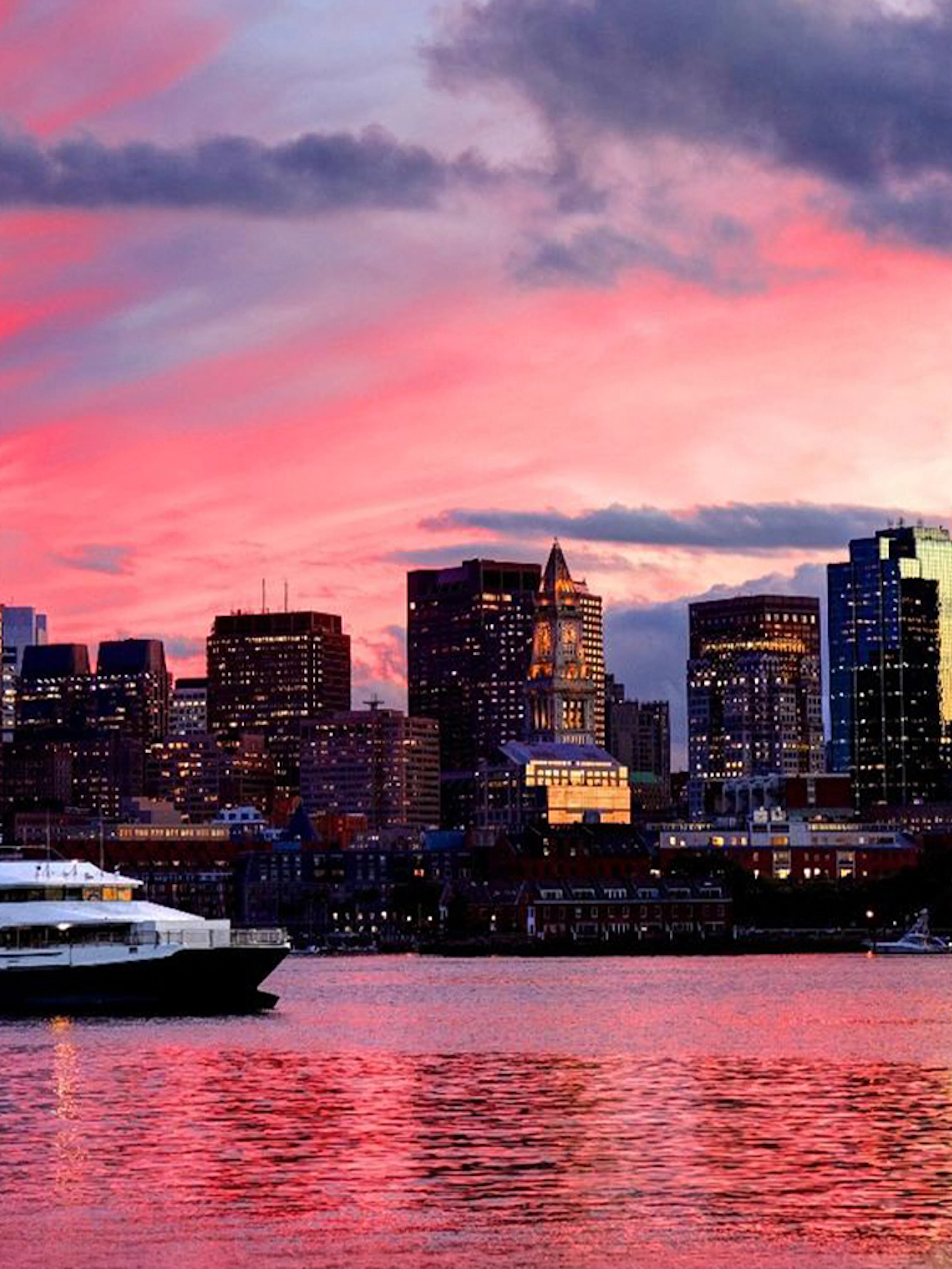 Cruise boat on Boston Harbor at sunset with city skyline in the background.