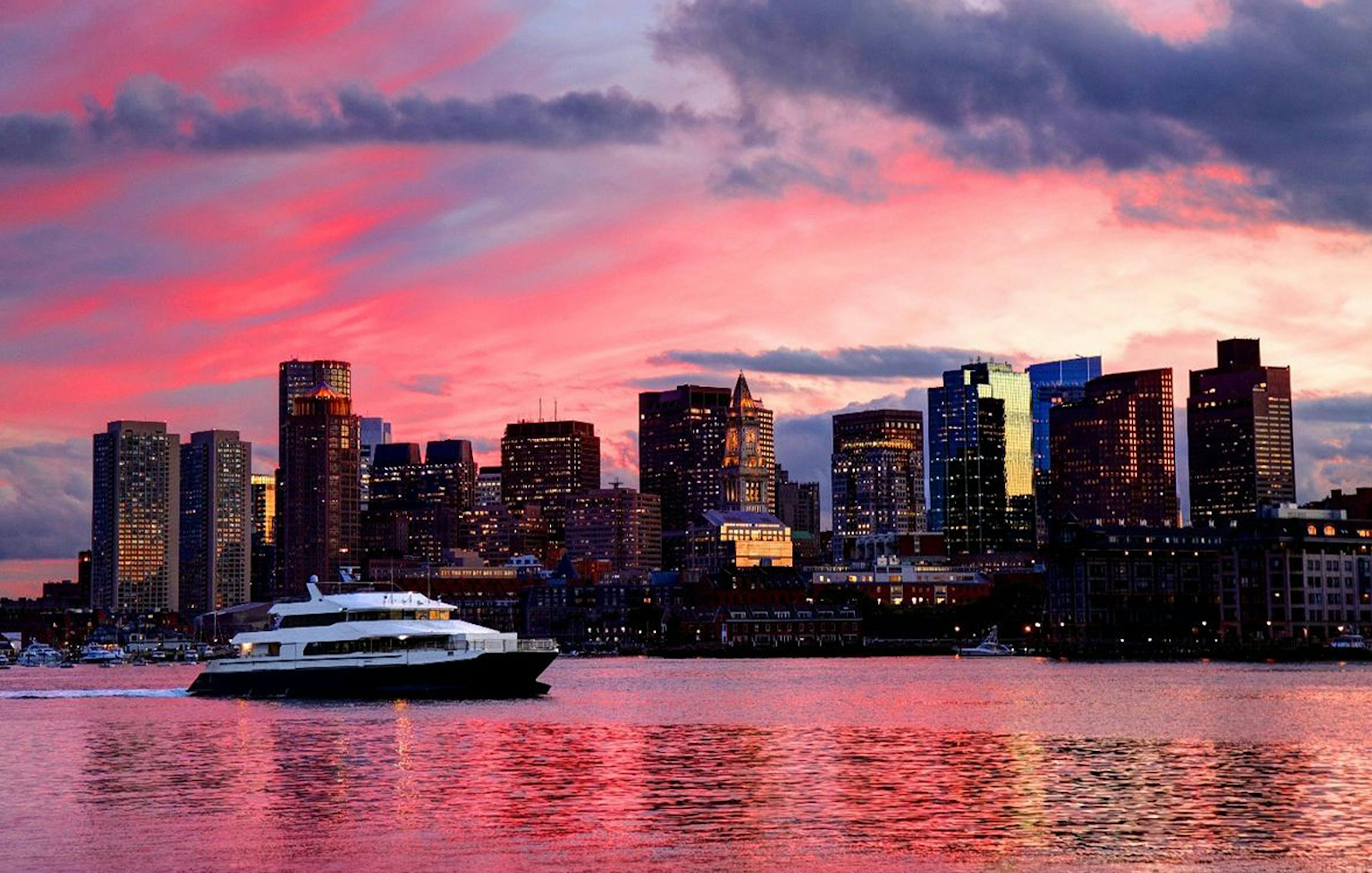 Cruise boat on Boston Harbor at sunset with city skyline in the background.