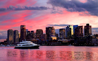 Cruise boat on Boston Harbor at sunset with city skyline in the background.