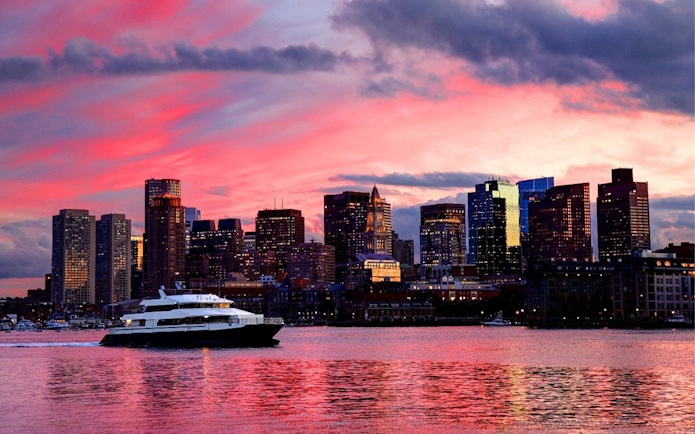 Cruise boat on Boston Harbor at sunset with city skyline in the background.