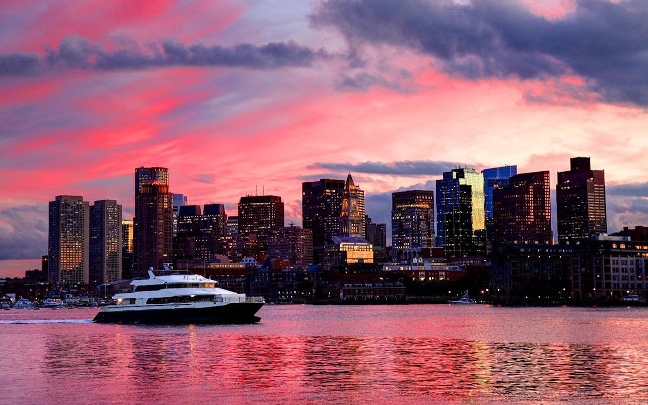 Cruise boat on Boston Harbor at sunset with city skyline in the background.