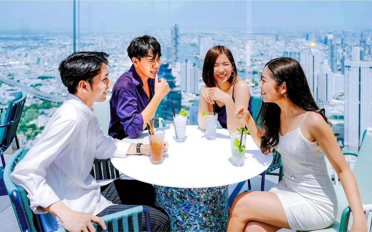 Friends enjoying drinks at Mahanakhon SkyWalk, Bangkok, with city skyline view.