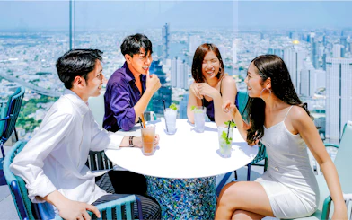 Friends enjoying drinks at Mahanakhon SkyWalk, Bangkok, with city skyline view.