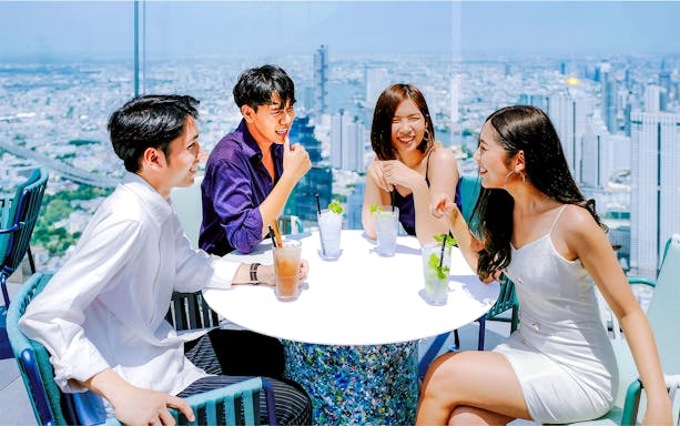 Friends enjoying drinks at Mahanakhon SkyWalk, Bangkok, with city skyline view.