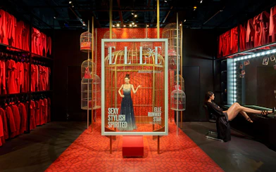 Fashion exhibit with model in a cage display, surrounded by red dresses and birdcages.