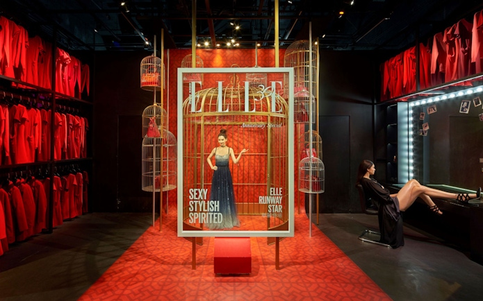 Fashion exhibit with model in a cage display, surrounded by red dresses and birdcages.