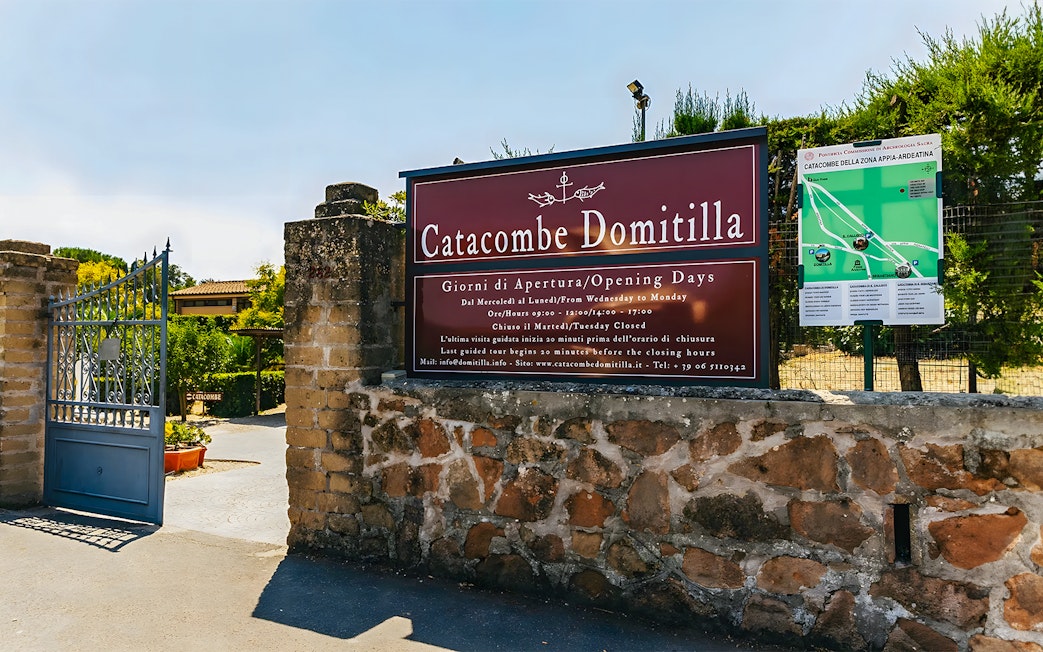 Entrance to Catacombs of Domitilla with sign displaying opening hours and map.