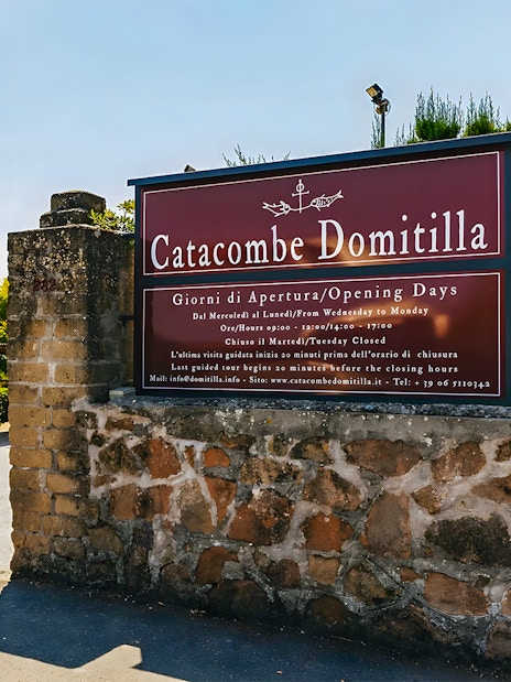 Entrance to Catacombs of Domitilla with sign displaying opening hours and map.