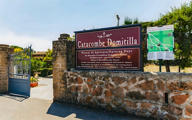 Entrance to Catacombs of Domitilla with sign displaying opening hours and map.