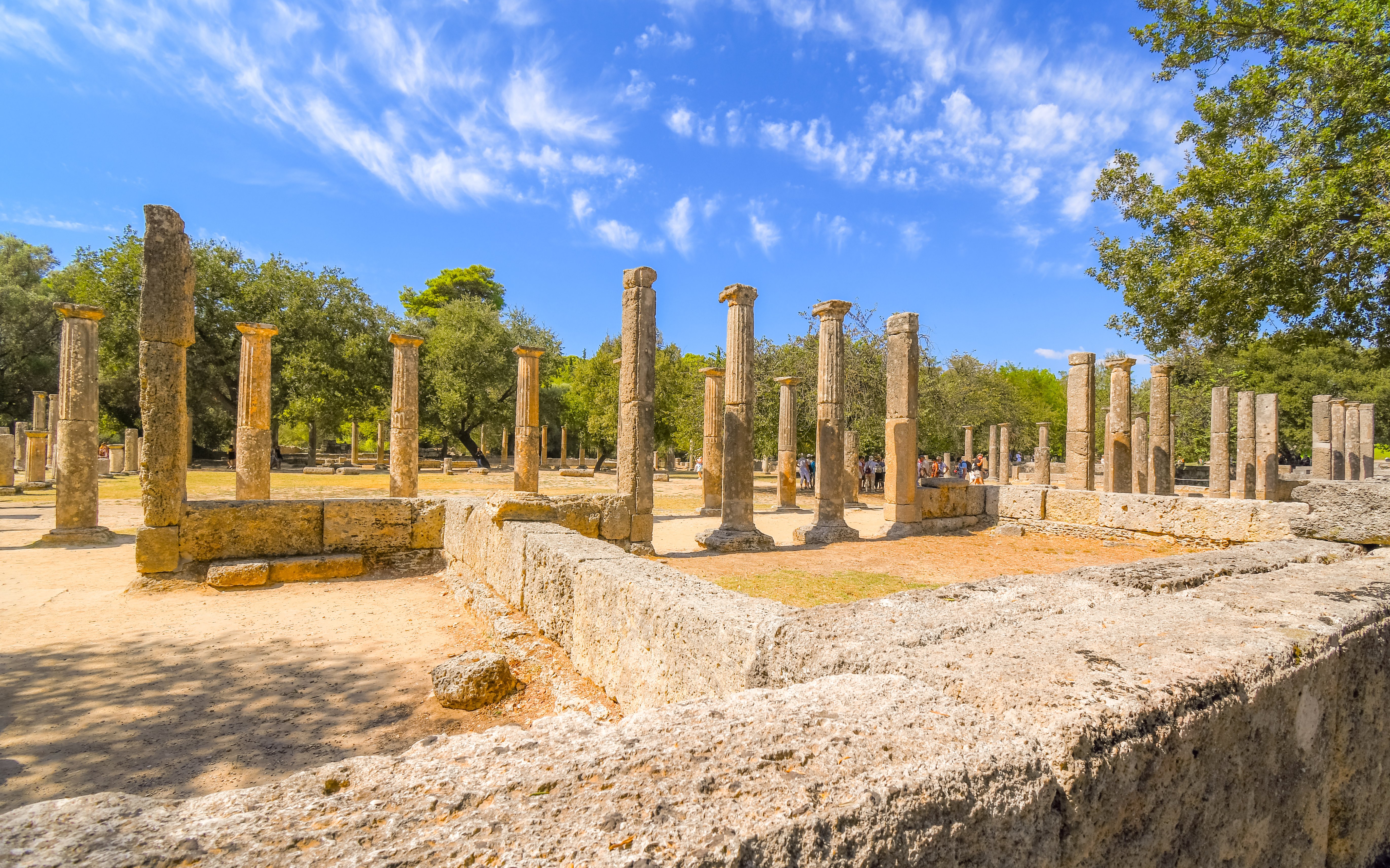 Ruins of the Palaestra wrestling school at ancient Olympia, Greece, with stone columns and trees.