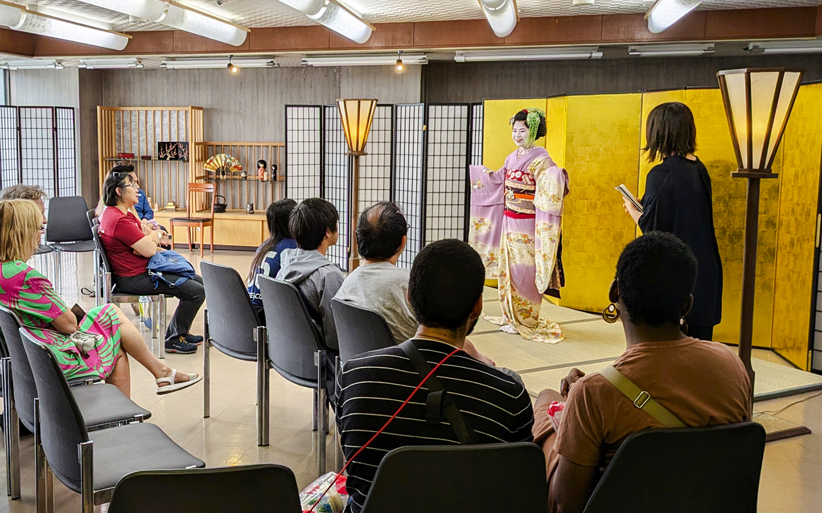 Maiko performing for seated audience in room with traditional Japanese screens.
