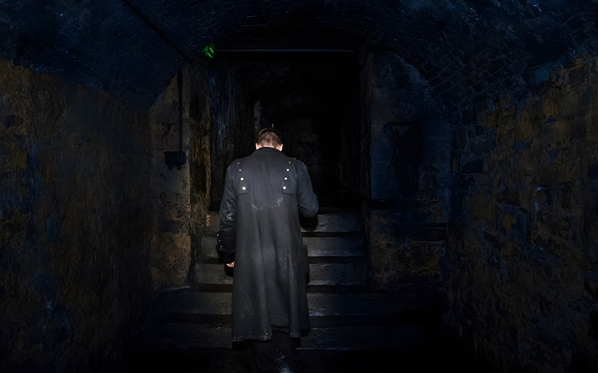 Man in dark coat ascending stone steps in dimly lit underground passage.