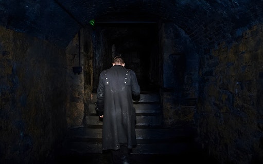 Man in dark coat ascending stone steps in dimly lit underground passage.