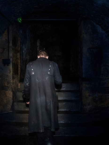 Man in dark coat ascending stone steps in dimly lit underground passage.