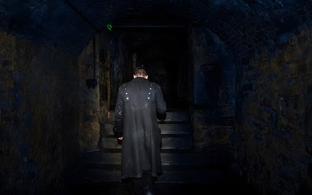 Man in dark coat ascending stone steps in dimly lit underground passage.