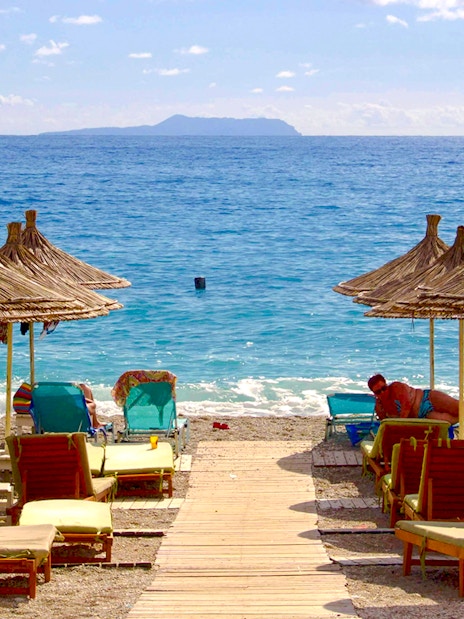 Sun loungers and straw umbrellas on Dhërmi beach, Albania, with clear blue sea.