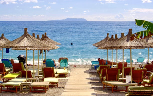 Sun loungers and straw umbrellas on Dhërmi beach, Albania, with clear blue sea.