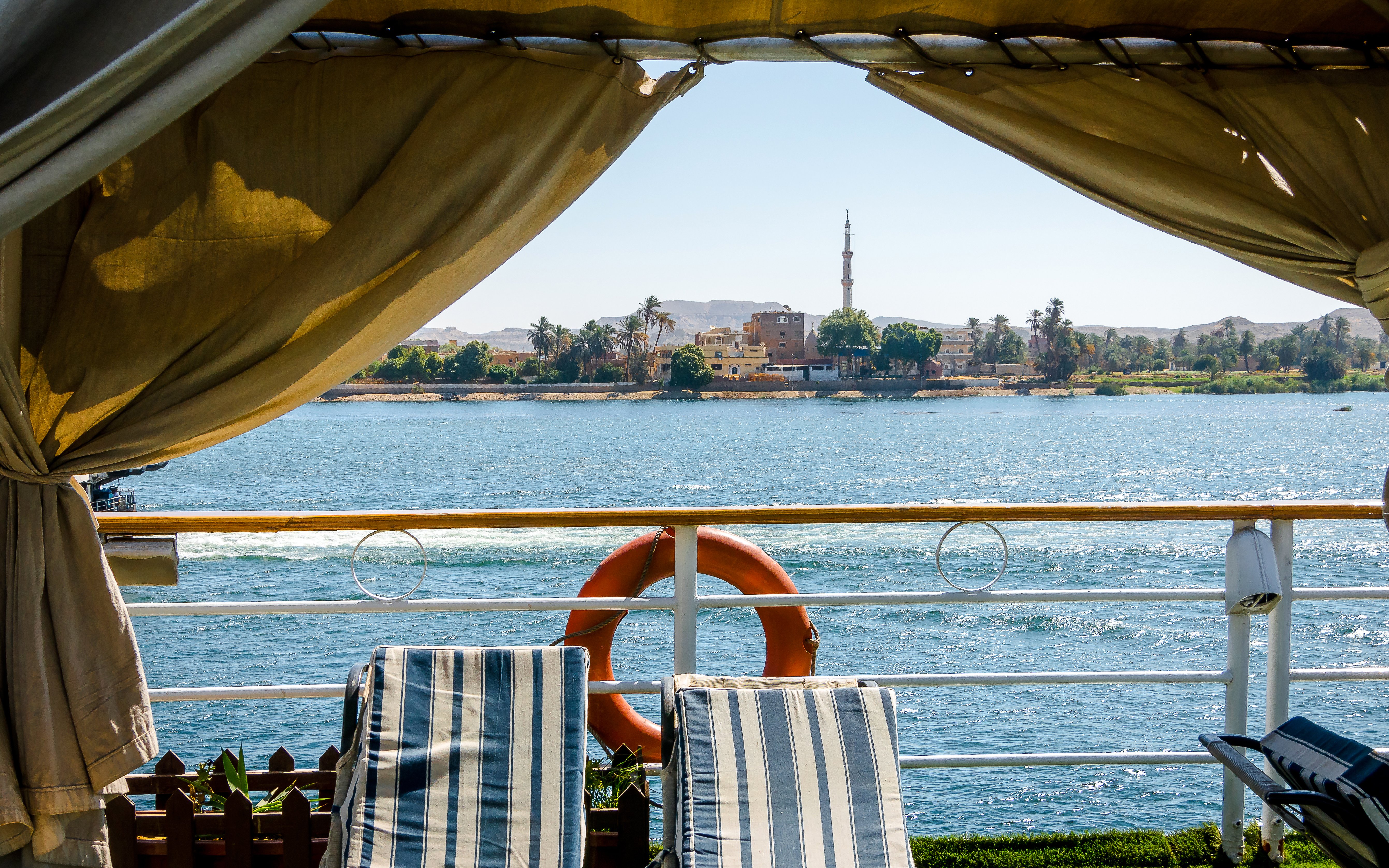 Sunlit view from Nile River cruise ship with deck chairs and distant minaret.
