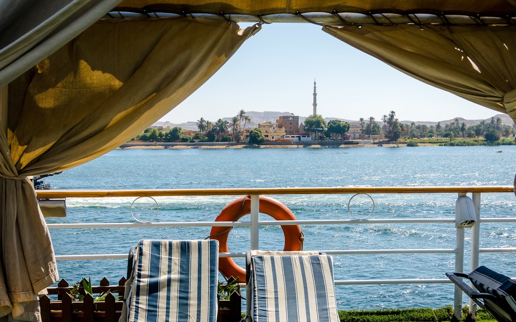 Sunlit view from Nile River cruise ship with deck chairs and distant minaret.