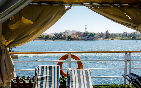 Sunlit view from Nile River cruise ship with deck chairs and distant minaret.