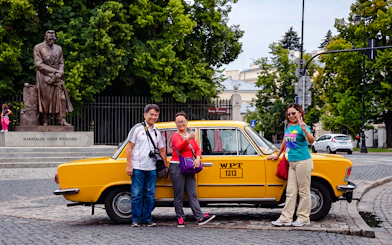 Tourists posing with a yellow Fiat 125p near the Józef Piłsudski statue in Warsaw.