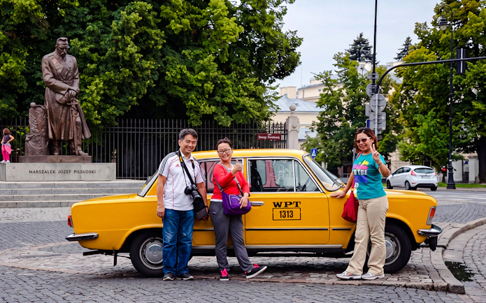 Tourists posing with a yellow Fiat 125p near the Józef Piłsudski statue in Warsaw.