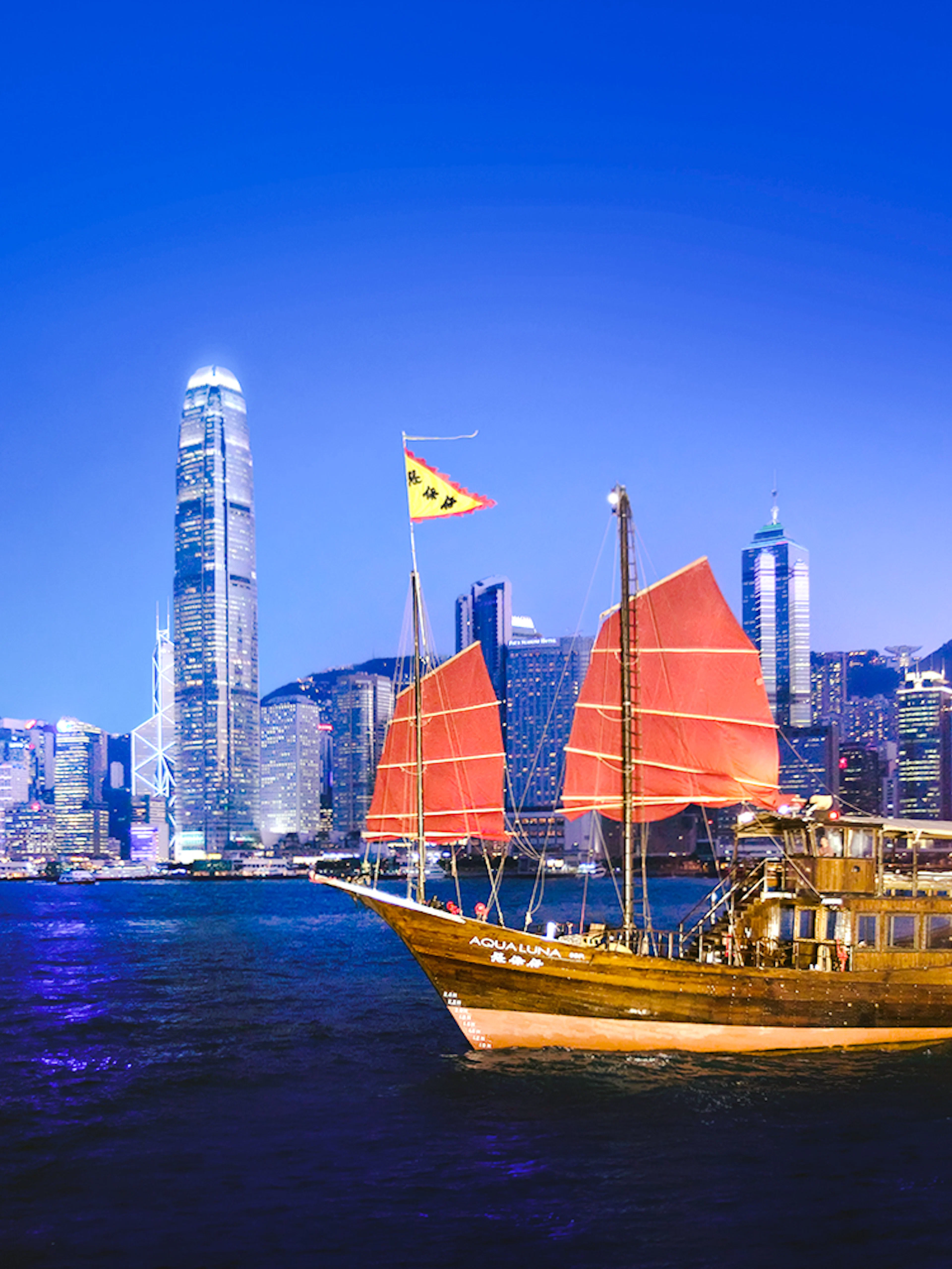 Traditional junk boat sailing in Victoria Harbour with Hong Kong skyline at dusk.