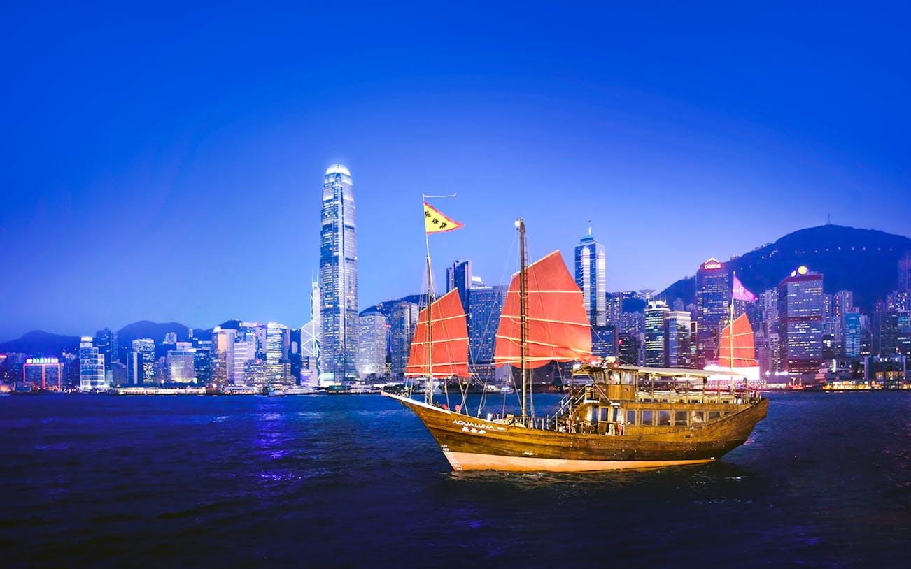 Traditional junk boat sailing in Victoria Harbour with Hong Kong skyline at dusk.