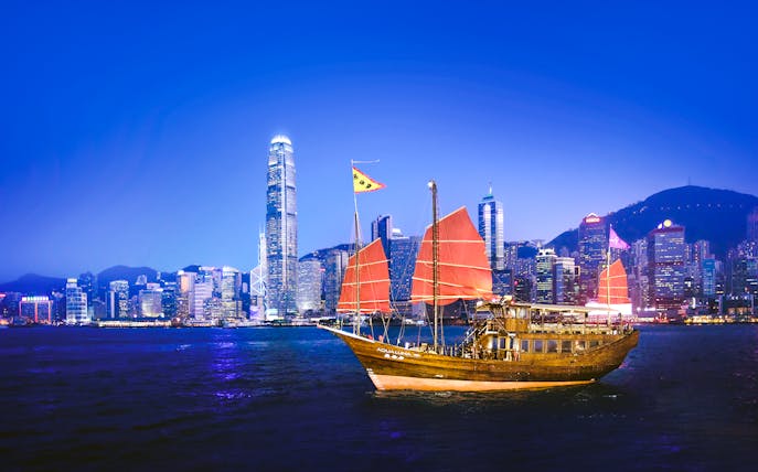 Traditional junk boat sailing in Victoria Harbour with Hong Kong skyline at dusk.
