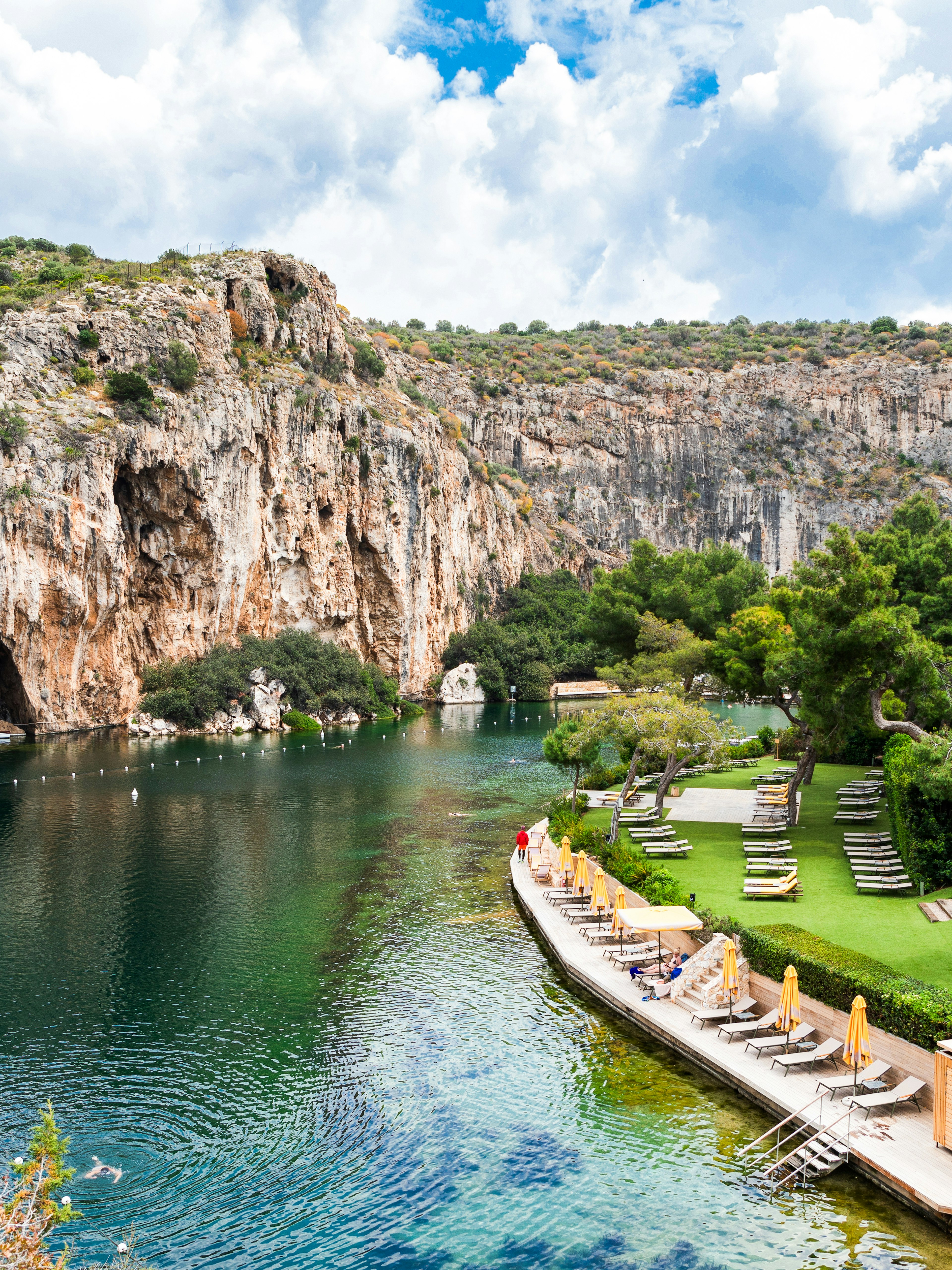 Sunbeds along the shore of Vouliagmeni Lake, Greece, with rocky cliffs and greenery.