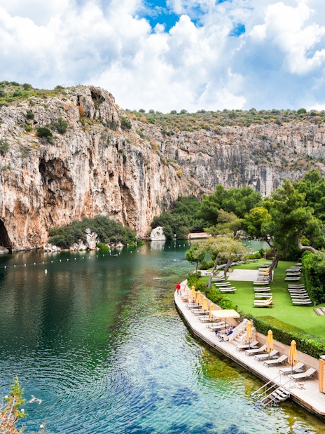 Sunbeds along the shore of Vouliagmeni Lake, Greece, with rocky cliffs and greenery.