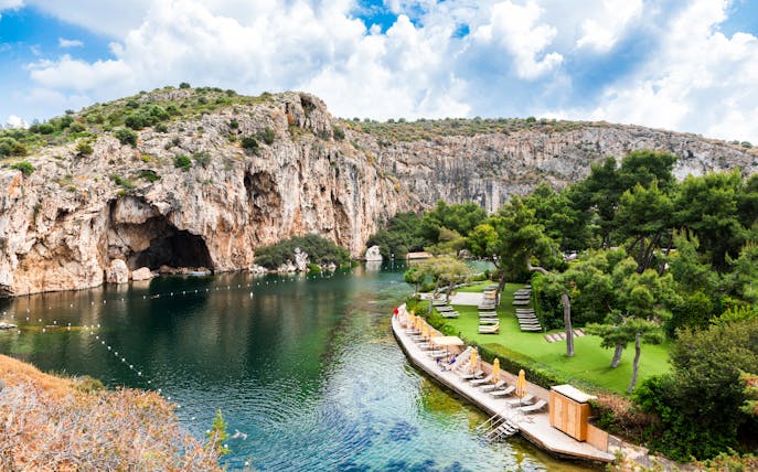 Sunbeds along the shore of Vouliagmeni Lake, Greece, with rocky cliffs and greenery.