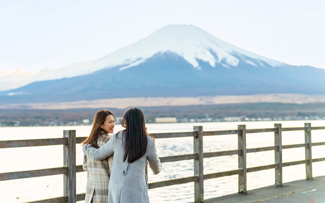 Two women walking on a lakeside path with Mount Fuji in the background.
