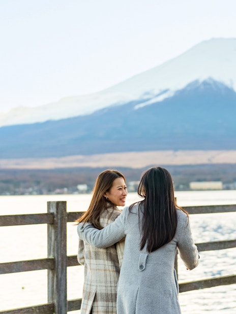 Two women walking on a lakeside path with Mount Fuji in the background.