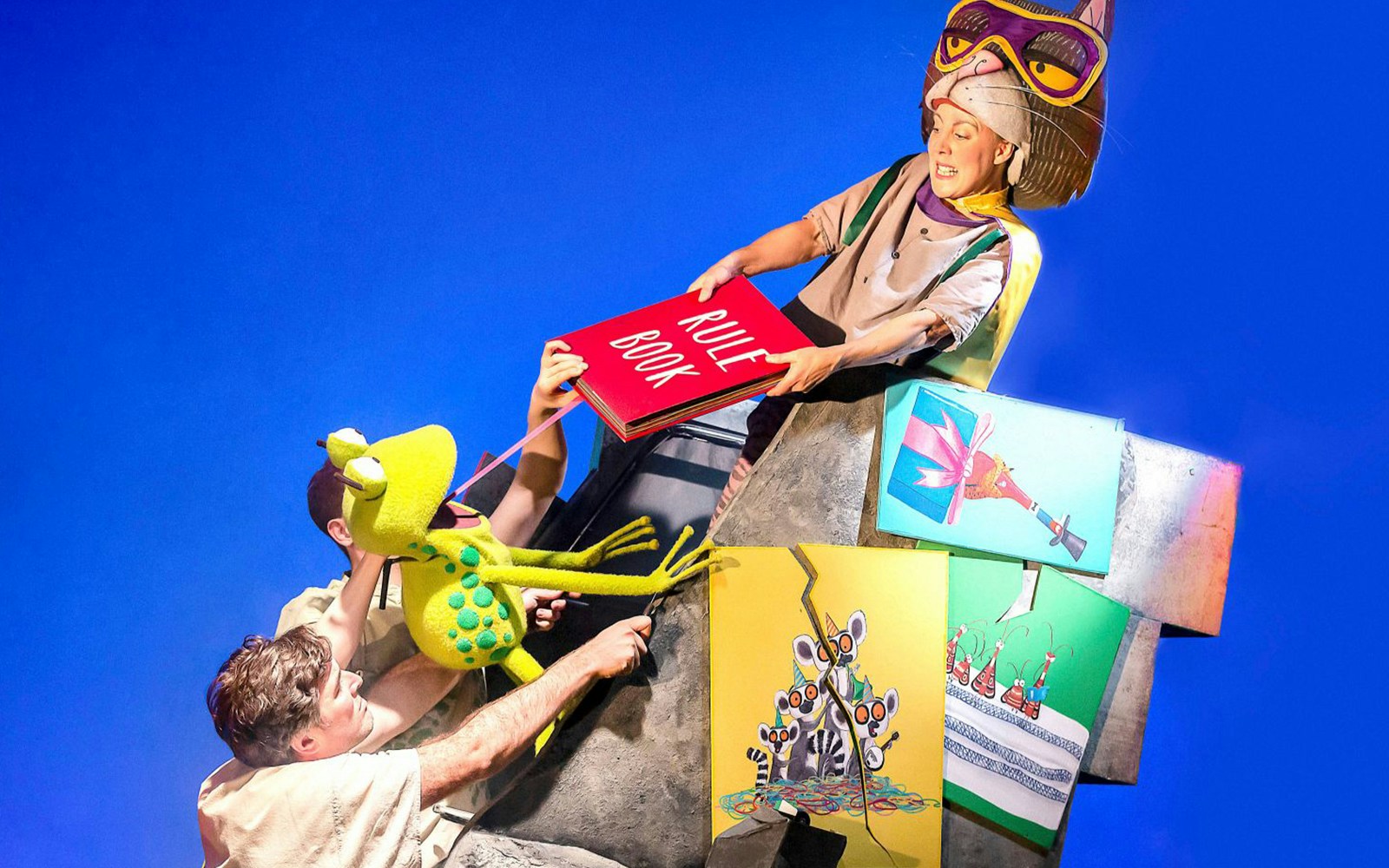 Actors performing "Oi Frog & Friends!" with a frog puppet and a large rule book on stage.