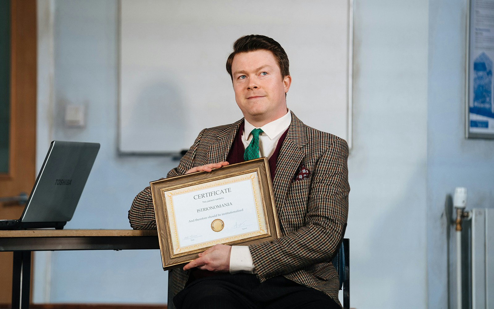 Man holding a framed certificate in a scene from "Accidental Death of an Anarchist" play.