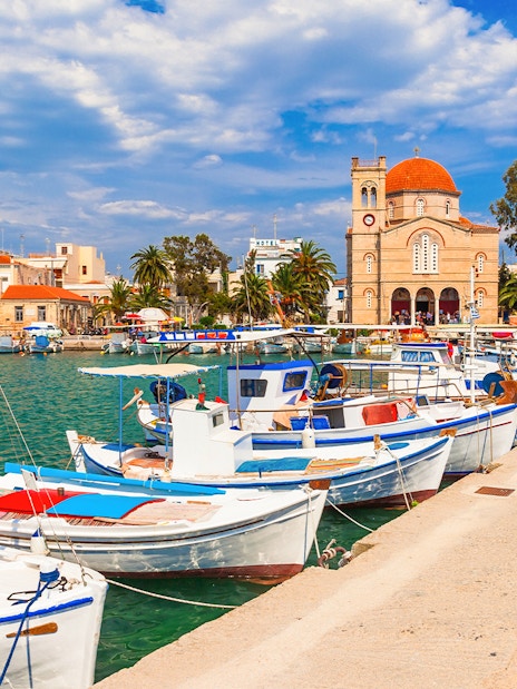 Boats docked at Aegina Island harbor with a church and buildings in the background, Greece.