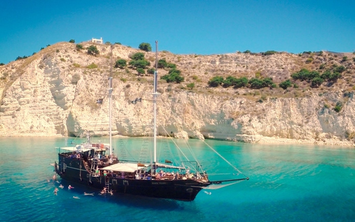 Pirate ship cruising in Souda Bay with swimmers near rocky cliffs.