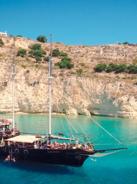 Pirate ship cruising in Souda Bay with swimmers near rocky cliffs.
