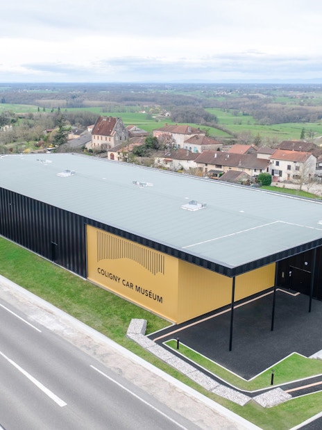 Coligny Car Museum building in Lyon, France, surrounded by countryside and village houses.