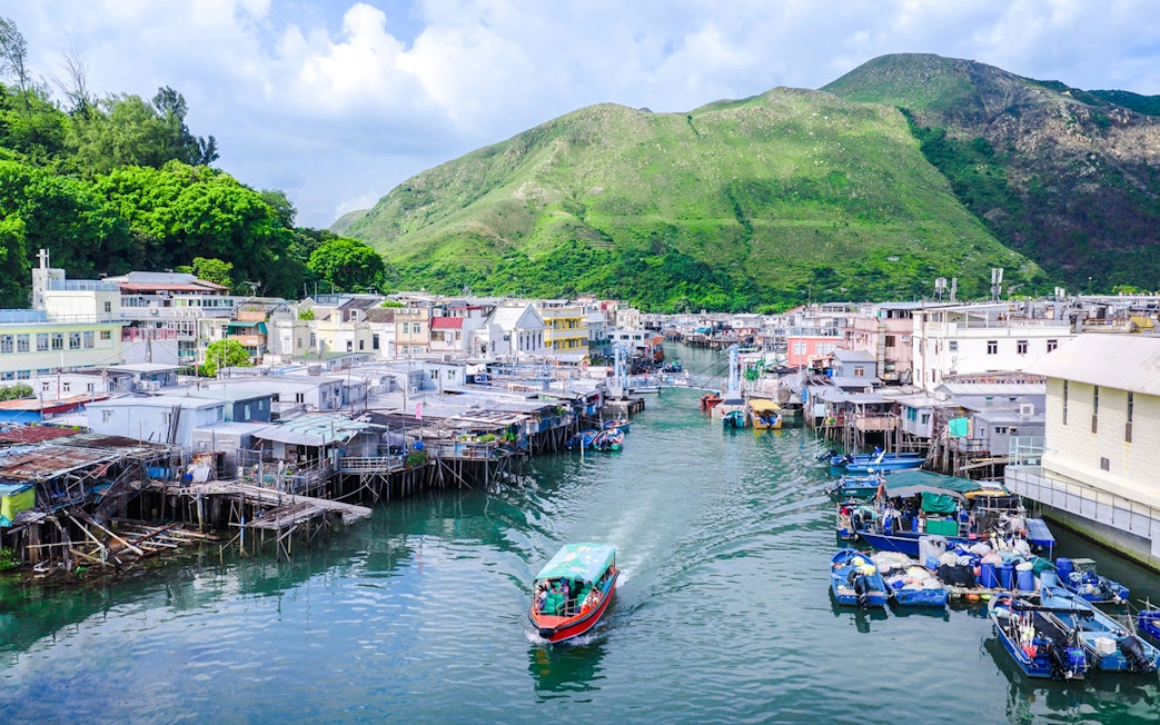 Boat navigating through Tai O fishing village with stilt houses and green hills in the background.