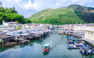 Boat navigating through Tai O fishing village with stilt houses and green hills in the background.