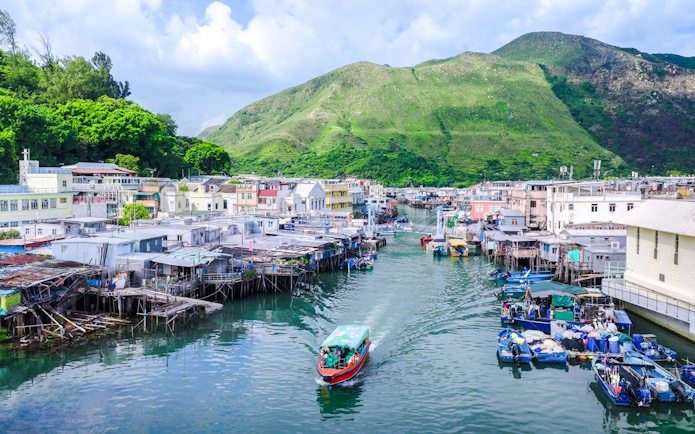 Boat navigating through Tai O fishing village with stilt houses and green hills in the background.